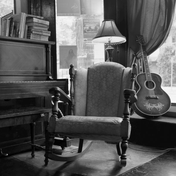 The interior of the Froth House, a coffee shop located in the Regent Street Neighborhood. A rocking chair is sitting in a corner, with a piano on the left. On the right is a guitar displayed in front of the window. There is a floor lamp behind the chair, and books are stacked on top of the piano.