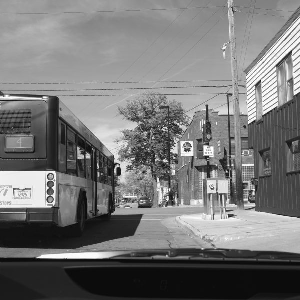 View through windshield of a car of the number four bus waiting at a traffic light on the corner of Williamson and Baldwin Streets. Cars are parked at the curb, and other cars are moving down the street. The Seton House Office is on the right at the corner. Across the street is the Crystal Corner Bar, with a sign advertising Pabst Blue Ribbon. 
