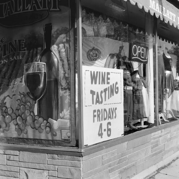 A sign in the window of Mallatt's Pharmacy and Costumes advertising wine tastings. The storefront window is decorated with Halloween costumes, such as Superman and Supergirl, and images of pumpkins, grapes, a wine glass and a wine bottle.