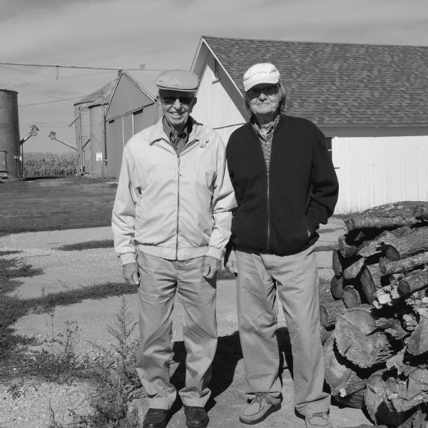 Two retired farmers, Richard Quinney, photographer, on the right wearing a baseball cap with the phrase "Life is good" stitched on it, and his cousin, wearing a flat cap on the left, are standing side by side next to a pile of firewood. Behind them are two barns and three small silos. A field of corn is in the far background. 