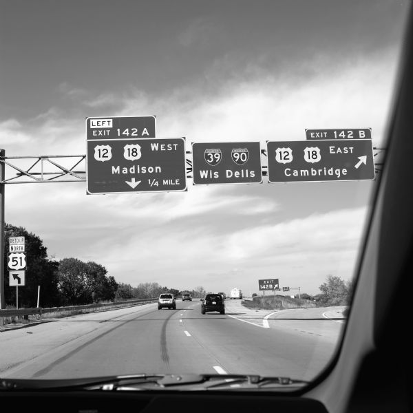 View through car windshield of road signs above Interstate 90. The exit for highway 12/18 towards Cambridge is on the right, and the exit for 12/18 towards Madison is on the left.
