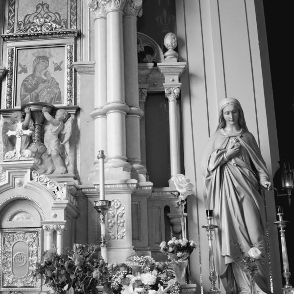 The interior of the Holy Redeemer Catholic Church on West Johnson Street. A statue of Mary is on the right between two large candle holders. There are roses in a group of vases in the foreground. On the left on the wall is a painting of Madonna and Child. Sculptures of two cherubs are holding up a pedestal underneath the painting.