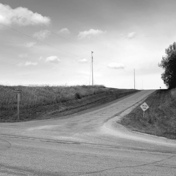 Two roads converge forming a crossroads. There are three utility poles standing in an open field of tall grass along Enchanted Valley Road which stretches into the distance. The road in the foreground going right to left is County Trunk K. A sign on the right reads: "Dead End." There are trees on the far right, with a mailbox silhouetted in the distance. 