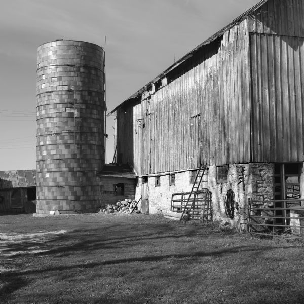 View across farmyard towards a silo standing in a corner made by two barn buildings. The barn is made of wood on a stone foundation, and is showing signs of wear. Lumber, fence sections, and a ladder are leaning against the barn. 