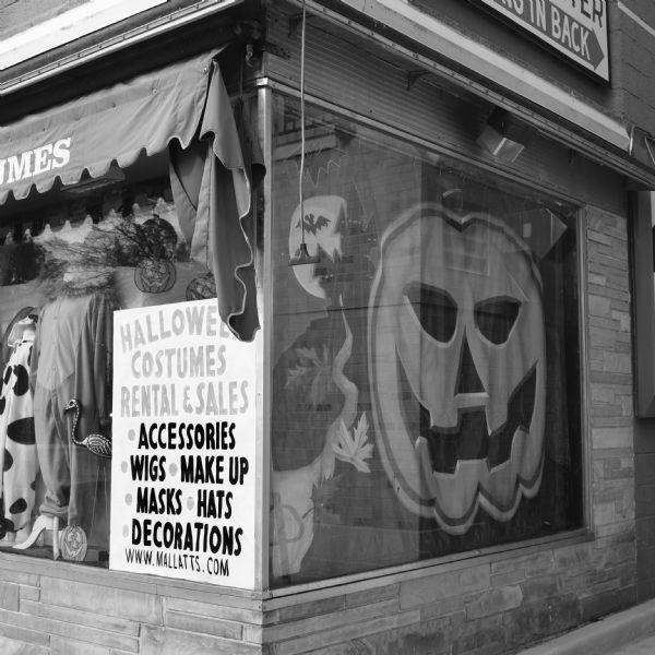 The corner storefront window of Mallatt's Pharmacy and Costumes on Monroe street is decorated with a Halloween display. Along the right side a cloth banner of a large jack-o'-lantern, bats, and a spooky castle fills up the entire window. Along the left, stickers of jack-o'-lanterns decorate the display of two onesies pajamas or costumes. A sign reads: "Halloween costumes rental & sales: accessories, wigs, make up, masks, hats, decorations. www.mallatts.com." 