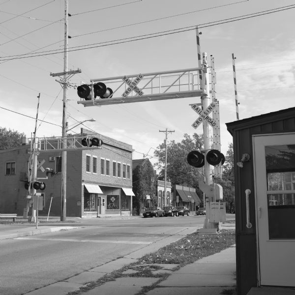 View from sidewalk down Lakeside Street towards a railroad crossing. Across the street beyond the railroad tracks are four buildings with cars parked in front of them. The first building on the left is the Cronometro bicycle shop, and the second building is Quintessence, an herb shop, which is covered in climbing vines. 