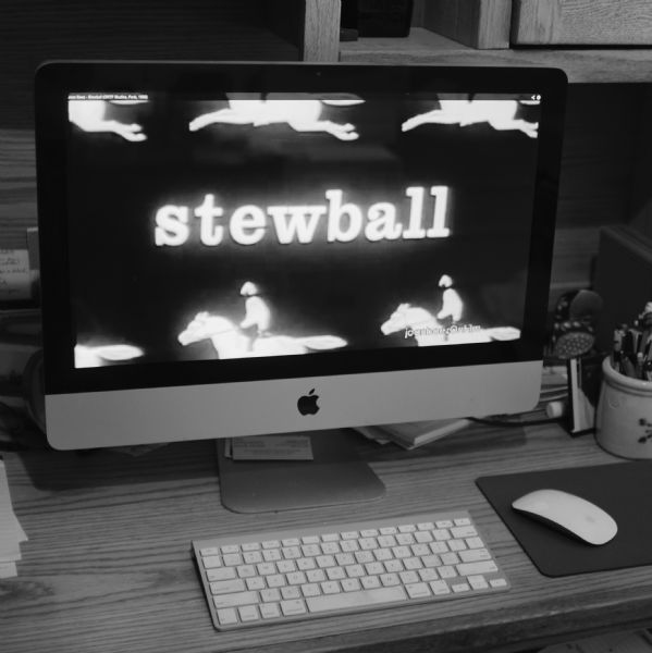Close-up view of a mac-monitor sitting on a wood desk with a wireless keyboard and mouse. A wooden cupboard is on the wall behind the monitor. A ceramic mug full of pens and pencils is on the desk to the right. On the monitor display is the word "stewball" along with six partial images of a jockey on a running horse. 