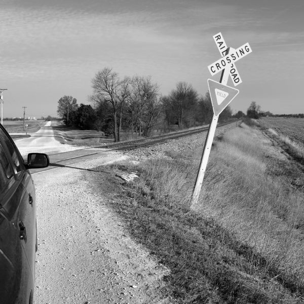 View along right side of car towards a railroad crossing at a gravel road in Dekalb County. The railroad tracks lead off to the right along a farm field. The gravel road is stretching to the horizon towards industrial buildings in the background.