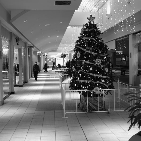 Christmas decorations adorn the hallway in a shopping mall. In the foreground is a Christmas tree on display inside a small fenced-in enclosure. Lights are suspended from the ceiling, and a wreath is hanging in the distance. There are a few people walking in the mall.