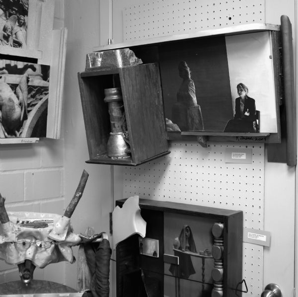 Postmodern works of art hanging, somewhat chaotically, on a corner wall. There are photographs of people combined with found object sculptures. Other sculptures are further down on the walls. 