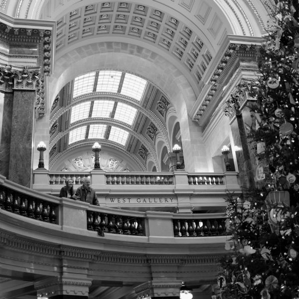 Interior view looking up in the Wisconsin State Capitol building. A tall tree decorated for Christmas is on the far right. Two people are standing on the circular balcony. Behind them on the next level up is another balcony, with the words: "West Gallery." On the ceiling are floral motifs and skylights.