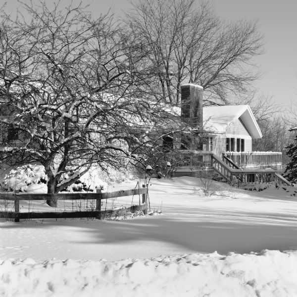 View across snow-covered ground, towards the photographer'shouse with a front deck. The winter landscape includes a wood fence and tree on the left.