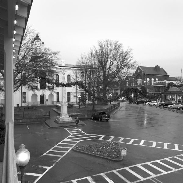 Elevated view of the Lafayette County Courthouse from the balcony of Square Books bookstore. Christmas lights, wreaths, and garlands decorate the square, courthouse, trees, and buildings across the street. The courthouse is a white building with a small clock tower and a balcony decorated with columns. In front of the courthouse is a tall pillar with a sculpture of a confederate soldier. A man is walking across the square. Cars are parked on the road road along the right side of the square near shopfronts.