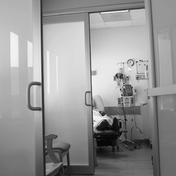View down hallway towards a person, obscured by a doorway, resting in a reclining hospital chair while receiving an infusion of chemicals for cancer treatment.