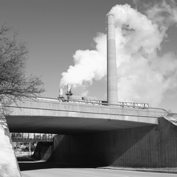 Smokestacks rising over a short underpass, billowing exhaust. Snow is covering the side of the pass and the ground on the other side.