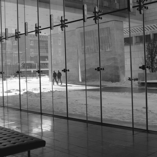 Indoor view through the lobby of the Chazen Museum towards glass walls which look out on East Campus Mall. Snow is on the ground, and two people are walking on the shoveled section between the museum and the Kohler Art Library underneath a skyway that connects the two buildings. Across the street is the School of Journalism and Mass Communication.