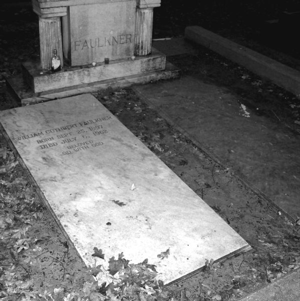 Gravestone of William Cuthbert Faulkner and his wife, Estelle Faulkner, in St. Peter's Cemetery. The markers and gravestones are weathered and dirty, and the ground around the stones is muddy and covered in leaves. A few tea candles and a bottle of whiskey are sitting under the carved marker which reads: "Faulkner." Estelle's gravestone is too dark to read the inscription. William's gravestone reads: "William Cuthbert Faulkner. Born Sept. 25, 1897. Died July 6, 1962. Beloved, Go With God."
