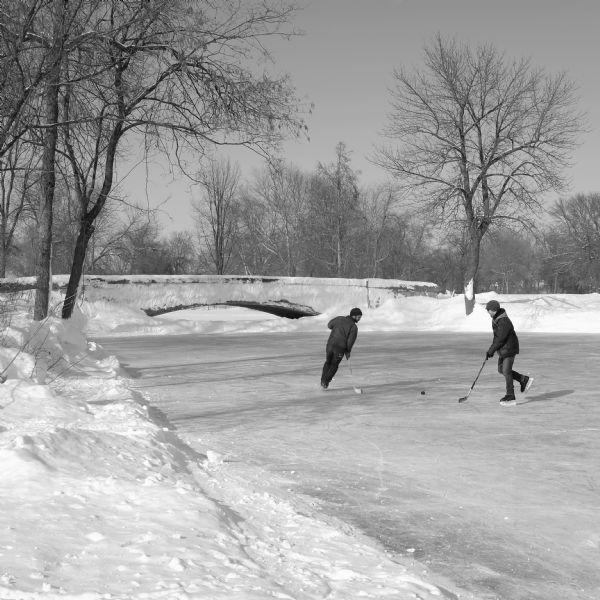 Two young men are playing hockey on the lagoon at Tenney Park. Snow is on the ground and the small foot bridge is in the background.