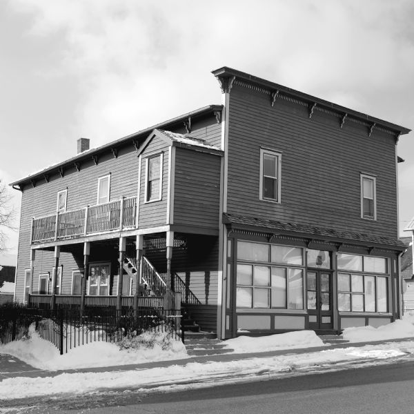 View across street towards the historic Blue Mounds Opera House. Exterior stairs lead to porches on the ground and second floor. A small metal fence surrounds the space on the left side of the building. Snow is on the ground. The ground floor in front has large windows and a double door.