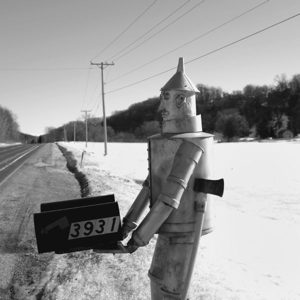 Close-up of a life-sized tin man sculpture with a hatchet attached to his side holding two mailboxes at the side of Highway 78. Snow is covering the ground, but the road is plowed. Trees are on the hill in the background on the right across a snowy field.