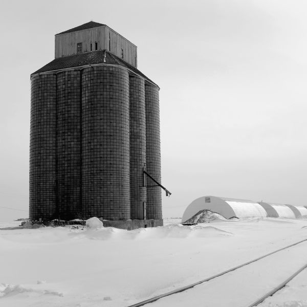 View across snow-covered ground towards a large grain elevator. Railroad tracks run diagonally in the foreground. Three arched storage sheds spaced slightly apart are in a row to the right of the grain elevator. 