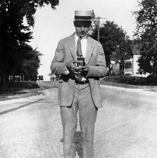 Portrait of a man wearing a suit, tie and boater hat standing in the middle of a cobbled road intersection looking down at a small folding camera he is holding in his hands. There are houses in the background on the right, and an automobile is on the crest of the hill in the far background. This photograph of Floyd Quinney comes from Richard Quinney's old family album.