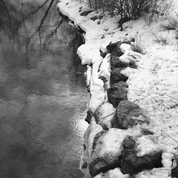 View looking down at the bank of the Yahara River. The sun is being reflected in the water near the snow covered stones on the shoreline.