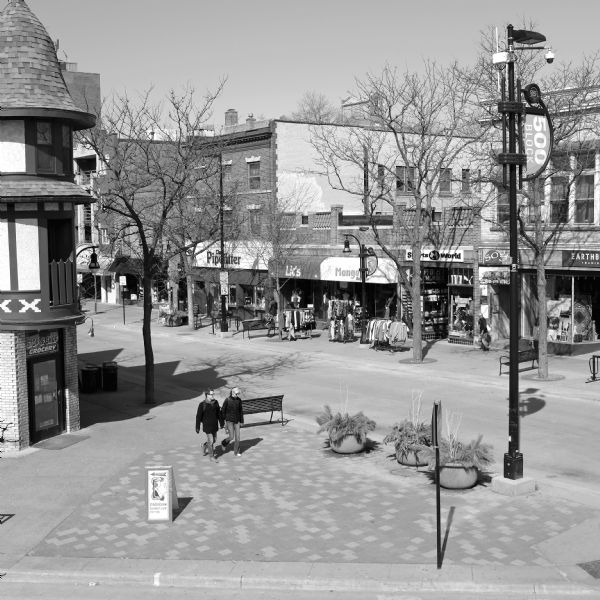 Elevated view of the corner of State Street and Gilman Street. A man and a woman are walking towards the corner away from the "Stop & Shop Grocery" behind them. A woman is sitting on a bench on the other side of the street near racks of clothing on display on the sidewalk.