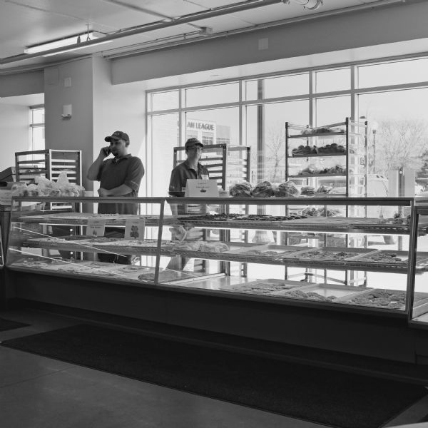 A man and a woman are standing behind the counter at Lane's Bakery. The glass display case has three rows of baked goods. There are three tall movable racks with trays of baked goods near the large windows with a view of cars in the parking lot. The Urban League of Greater Madison building is in the background on the left.