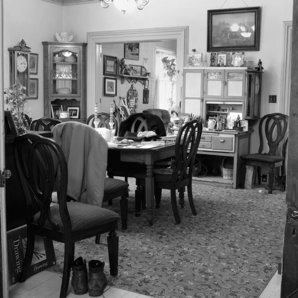 Interior view of a dining room full of knickknacks, furniture, clocks, a hutch and a cabinet. Coats are hanging on the back of the wooden chairs at the wood table. On the table are binders, papers, two candlesticks and a decorative tree holding Easter ornaments. Paintings and pictures are hanging on the wall. There is an open doorway leading into another room with a window.
