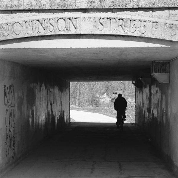 Silhouette of a person riding a bicycle through a pedestrian tunnel under East Johnson Street near the Yahara River and Tenney Park. Above the entrance to the tunnel is a carved sign that reads: "Johnson Street."