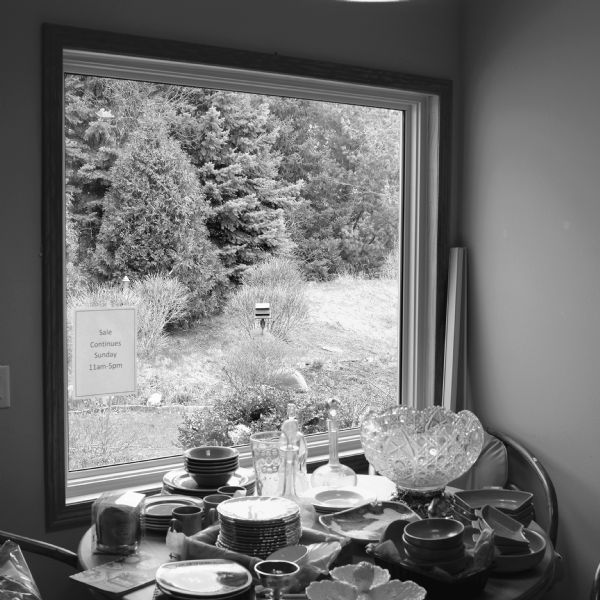 View of a table near a window displaying tableware, including dish sets, mugs, and a crystal bowl. The view out the window is of pine trees, shrubs and a bird feeder in the yard. A sign on the inside of the window reads: "Sale continues Sunday 11am-5pm."
