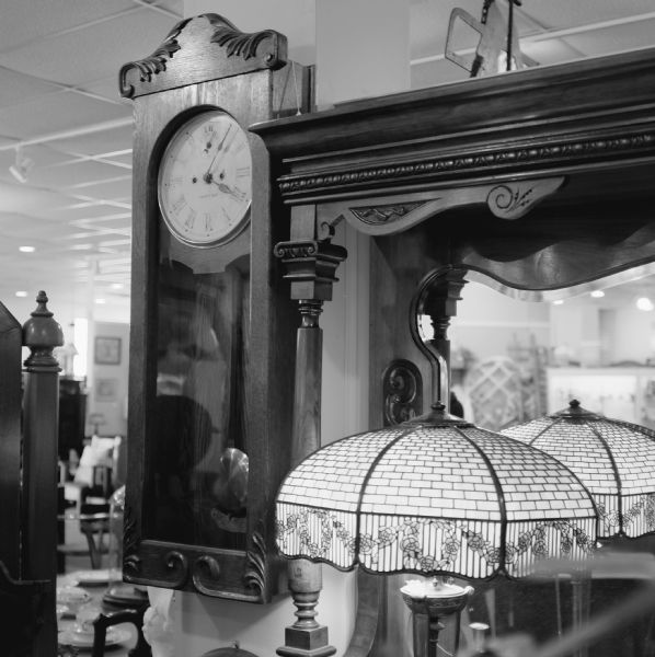 Oak-encased pendulum wall clock hanging in an antique store. In the foreground is a desk lamp, with a Tiffany style glass lamp shade with a rose motif, sitting on a ornately carved wooden dressing table with a mirror. Furniture, dishware, and other items are in the background on the left.
