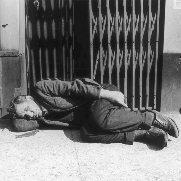 View of a man sleeping on the sidewalk near the side of a building in front of a folding steel door. 