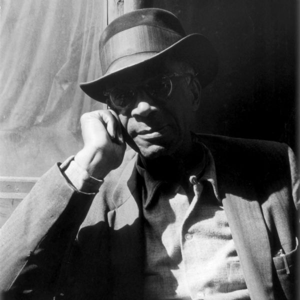 Waist-up portrait of an elderly man sitting outdoors. He is wearing glasses, a hat, a suit jacket, and buttoned shirt. He is smiling slightly and resting his cheek on his right hand.