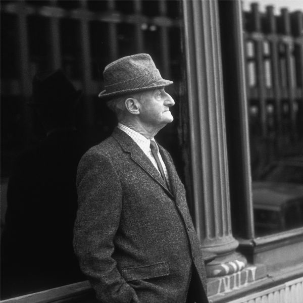 Profile view of a man dressed in a suit and wearing a hat. He is standing in front of a large picture window, which is reflecting the framework during construction of the World Trade Center.
