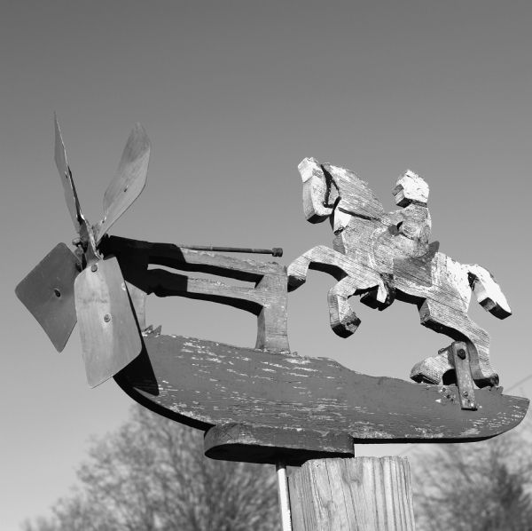 Close-up view of a decrepit wooden whirligig about to be retired. The paint on the figure of a man riding a horse is faded, and the blades of the whirligig are slightly warped.