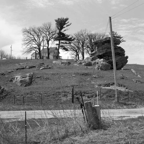 View across fence and road towards a house on a hill of outcroppings in the Driftless Area. There are fence lines, telephone and electric poles, and a few trees.