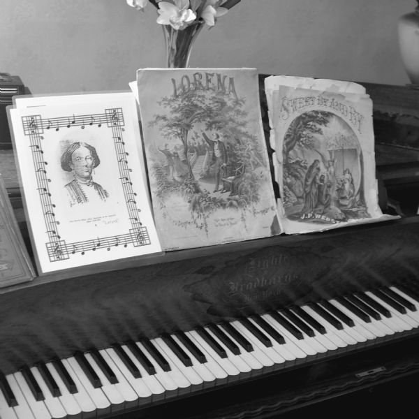 Close-up of a grand piano and old sheets of music in the historical house of the musician Joseph P. Webster. A vase of flowers is on top of the piano behind the sheet music. The two pieces of sheet music on the piano desk stand have the titles: "Lorena" and "Sweet By and By."