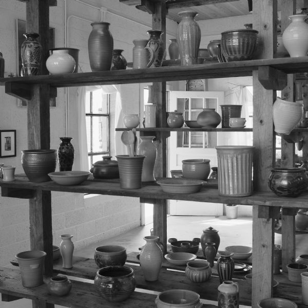 Display in a gallery of handmade ceramic bowls, vases, and vessels on open wooden shelves. Behind the shelves, pictures are displayed on the walls, and there are two windows and an open door.