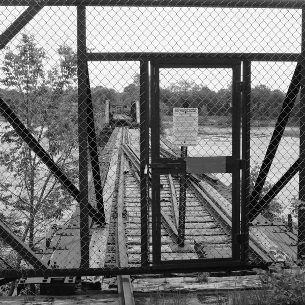 View through chain-link fence towards the railroad trestle that spans the Wisconsin River, worn with age, and split apart in the middle. There is a padlocked door in the fence, and just beyond is a sign that reads: "Wisconsin & Southern Railroad, No Trespassing."