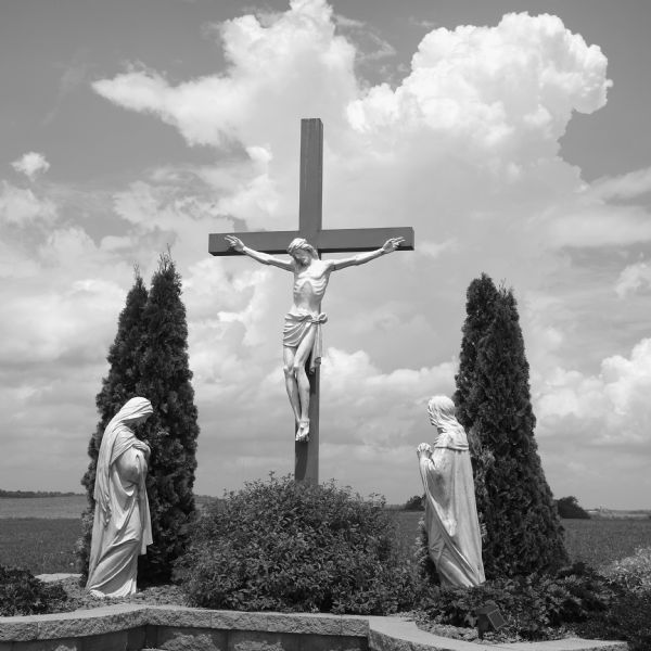 Three statues among plants and small trees in a cemetery representing the Crucifixion of Jesus. Two statues of women, probably Mary and Mary Magdalene, are on either side of the cross. In the background are newly planted fields of crops.