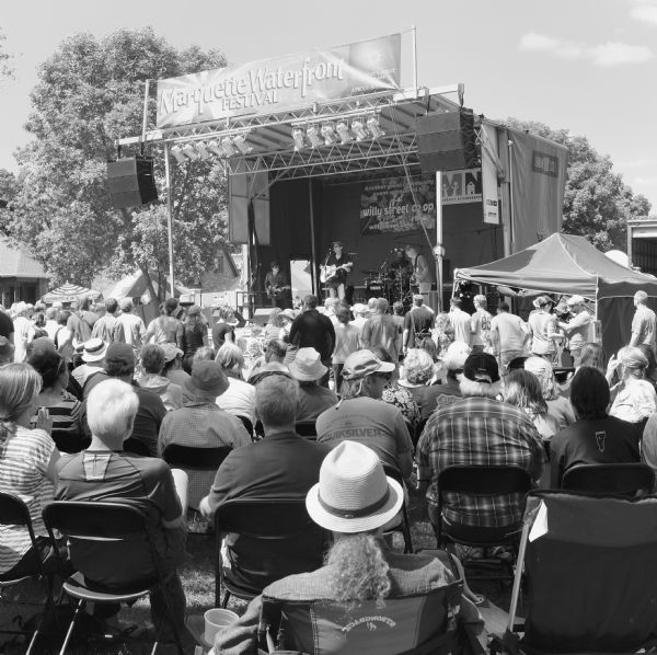 View from behind of an audience of men and women standing, or sitting in folding chairs, in front of a large stage. Four men are playing guitars, bass, and drums on the stage, and singing into microphones. Smaller tents surround the stage. A large sign above the bandstand reads: "Marquette Waterfront Festival."
