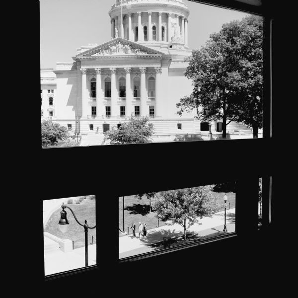 View of the capital and the square through a window on the top floor of the Wisconsin Historical Society Museum. Three people are walking on the sidewalk around the square.