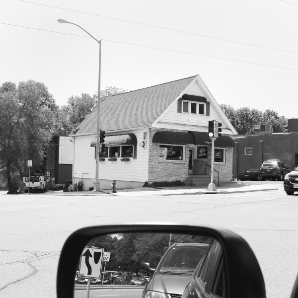 View from automobile across an intersection towards the Village Bar at the corner of Mineral Point Road and Glenway Street. Cars are waiting at a street light or are parked in the small parking lot. In the foreground, a driver's side mirror is reflecting more cars waiting for the green light, as well as golf carts at the Glenway Golf Course.