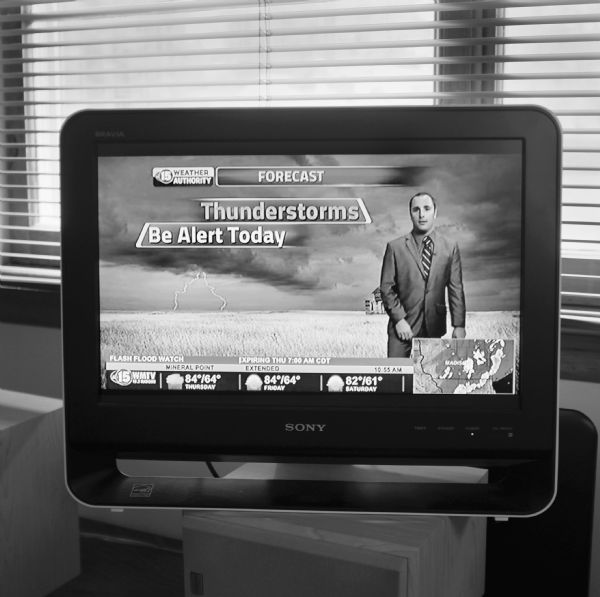 A Sony T.V. is sitting on top of a speaker in front of two windows with window blinds. On the screen, a man wearing a suit is standing in front of a screenshot of lightning striking a crop field. The weather forecast is for thunderstorms and a flash flood watch.