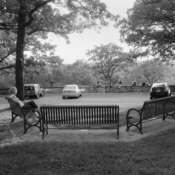 View of a woman sitting on one of three benches at the Sunset Point Overlook in Hoyt Park. In the background three cars are parked facing the stone wall of the Owen Parkway. Four people are sitting on the wall with their bicycles nearby. 