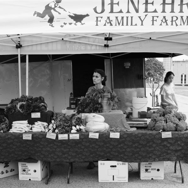 View at farmers' market of two women selling vegetables from behind a booth. The sign on their tent reads: "JenEhr Family Farm," and includes an illustrated image of a boy chasing after a hen and three chicks.