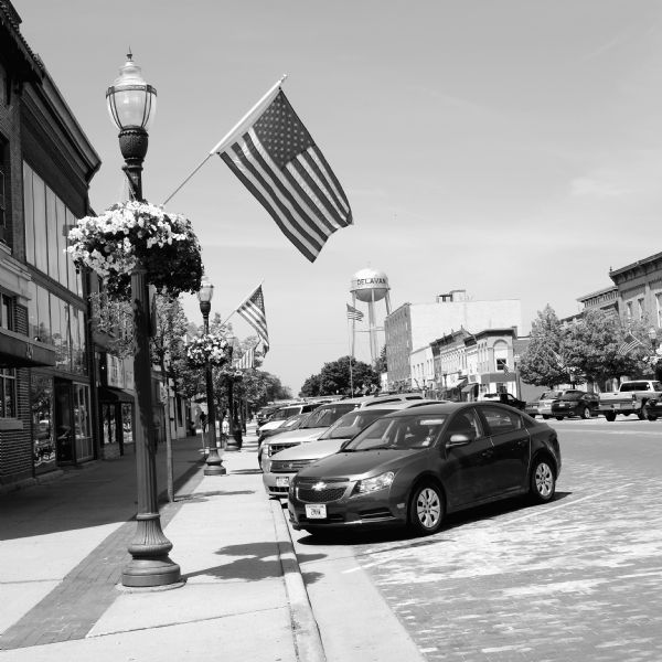 View down sidewalk towards cars parked diagonally at the curb, with a row of storefronts on the left. Along the sidewalk are lampposts, each decorated with a hanging basket of flowers and an American Flag. In the distance is flag pole near the Delavan water tower.