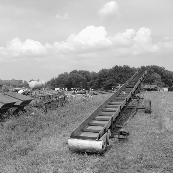 Farm equipment and machinery, including a corn elevator in the foregroung, stored in a grassy field. Cows are grazing in the background.